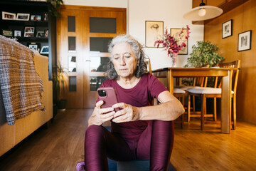 Woman in sportswear sitting on an exercise mat, checking her phone in the living room