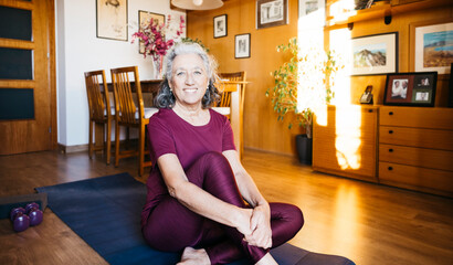 Senior woman sitting on yoga mat in living room, smiling at camera