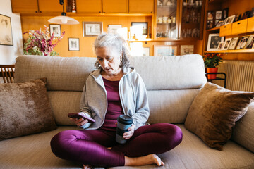 Mature woman sitting cross-legged at home wearing sport clothes, holding a phone and a reusable coffee cup after workout at home