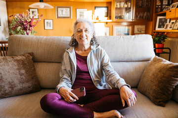 Senior woman relaxing and smiling on a couch, wearing sport clothes holding a smartphone after workout at home