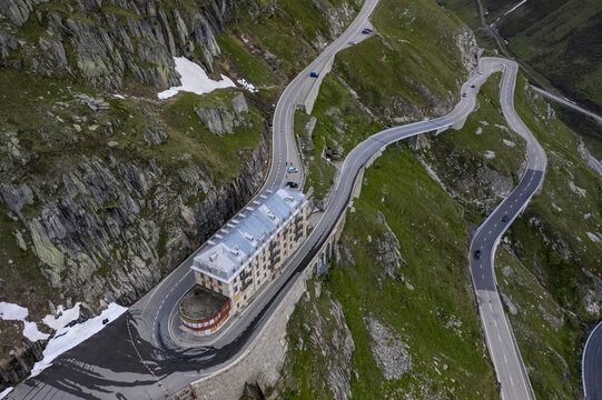 Aerial view of a building nestled beside a winding road snaking through rugged, green mountains, a blend of man-made structure and natural beauty, Obergoms, Valais, Switzerland.