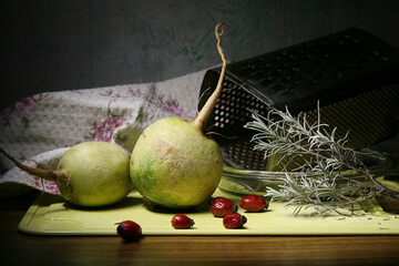 Two white radishes lie on a cutting board against a backdrop of a kitchen towel and an old grater.