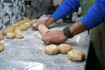 Making unleavened flatbreads. A man's hands roll out roti dough with a rolling pin. The surface is dusted with multigrain flour. Several balls of dough lie nearby. A gas stove is in the background.