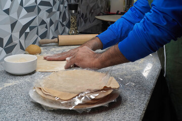 Making unleavened flatbreads. A man's hands touch a flatbread. Multigrain flour, a ball of dough and a rolling pin lie nearby. A stack of flatbreads is ready to freeze. A gas stove with bread baking.