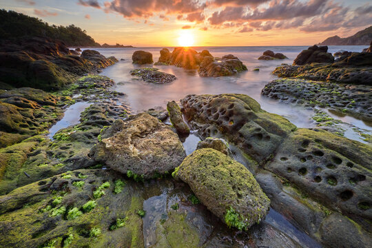 View of the sun's golden rays bursting over the horizon, illuminating the rugged, moss-covered rocks and tranquil waters of Wediombo Beach, Yogyakarta, Special Region of Yogyakarta, Indonesia.