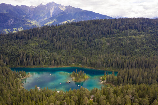 Aerial view of the crystal-clear turquoise waters of Lake Cauma surrounded by dense green forests and towering mountains under a cloudy sky, Flims, Grisons, Switzerland.
