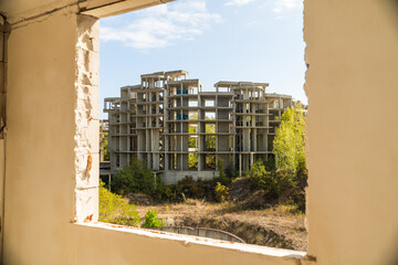 View of an unfinished concrete structure stands amidst greenery and a pit, framed by a weathered window, hinting at abandonment, Costa del croco, Tsarevo, Bulgaria.