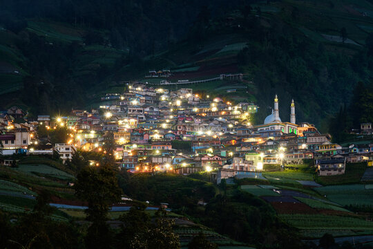 View of a village nestled in rolling green hills, dotted with twinkling lights and a prominent mosque, creating a serene night scene, Magelang, Central Java, Indonesia.