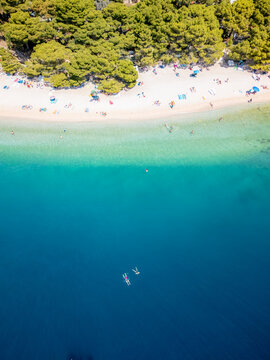 Aerial view of azure waters meet the sandy shore, lush trees fringe the coastline as tiny figures dot the beach, Brela, Split-Dalmatia County, Croatia.