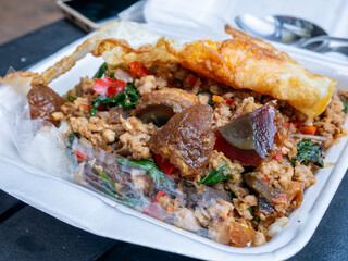 Traditional Thai street food, 'Pad Kra Pao Moo Sub Kai Yeow Ma' (Stir-fried minced pork with basil and preserved egg) served with rice and a fried egg in a foam box.