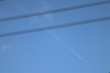 Jet plane flying high leaving white contrail in blue sky behind blurred power lines