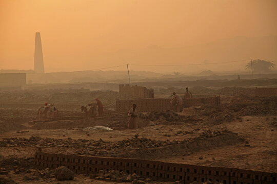 View of workers amidst the dusty landscape constructing brick structures under a hazy sky, with a towering chimney looming in the distance, Mithi, Sindh, Pakistan.