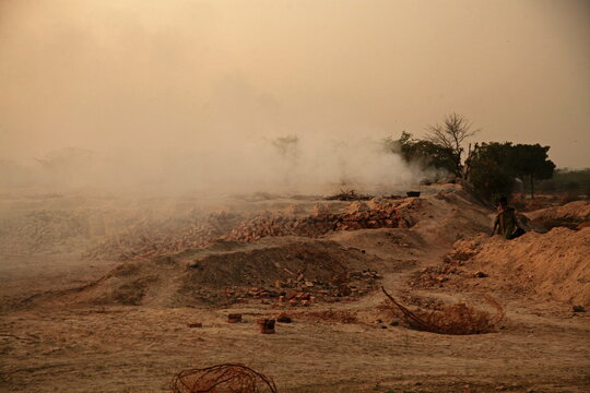 View of smoke billowing over brick kilns casting a haze across the arid landscape, where a lone figure works amidst the earthy tones, Mithi, Sindh, Pakistan.