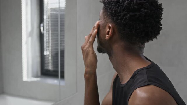 Tilt up view of Black young man applying hydrating cream to face when taking care of skin during morning routine