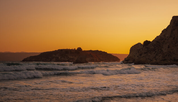 Dynamic ocean surf hitting rocky shoreline against dark cliff face at tranquil coastal sunset moment