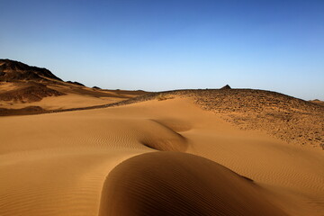 View of endless golden dunes rippling under a clear blue sky, with rocky outcrops puncturing the horizon in Noshki, Balochistan, Pakistan.