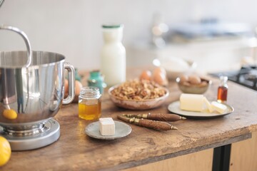 A sunlit kitchen counter holds baking essentials. A mixer stands ready, surrounded by butter, walnuts, eggs, milk, and honey, promising a delectable treat is about to be created