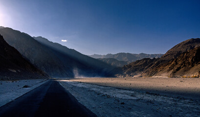 View of the long dark road cutting through a desolate landscape where sunlight pierces the high, jagged mountains, creating a stark contrast of light and shadow, Khaplu, Gilgit Baltistan, Pakistan.