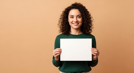 Cheerful young woman with curly hair holding a blank white poster board against a tan background, smiling and looking at the camera with copy space