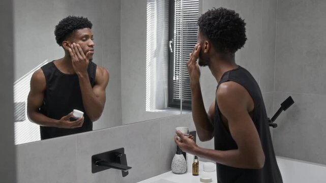 African American gen Z man applying face cream to his cheeks looking in mirror during self-care routine in morning