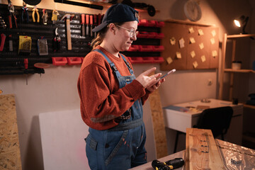 Female carpenter in overalls using smartphone for online shopping and payment in workshop