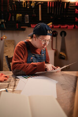 Female craftswoman studying new project details and reading paper blueprint in small workshop