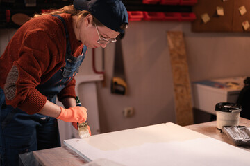Female artisan painting a wooden table with white paint in a small garage workshop. Woman restoring old furniture. DIY home improvement and furniture renovation