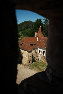 View of Bethlen Castle through a stone window, revealing its red-tiled roof and weathered walls against a backdrop of lush green hills, Cris, Transilvania, Romania.
