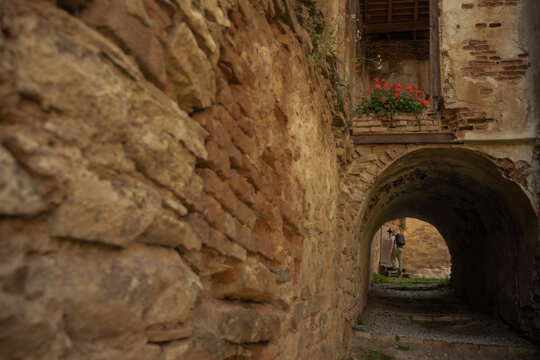 View of aged stone walls and a shadowed archway beckon towards a figure in the distance, framed by vibrant red flowers in a balcony, Cris, Transilvania, Romania.