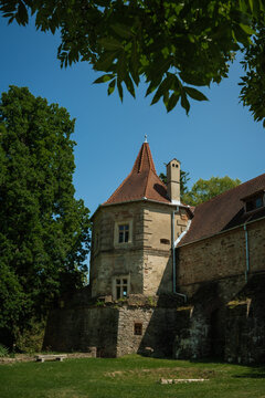 View of a historic stone castle tower under a bright blue sky framed by leafy branches and lush trees in Cris, Transilvania, Romania.