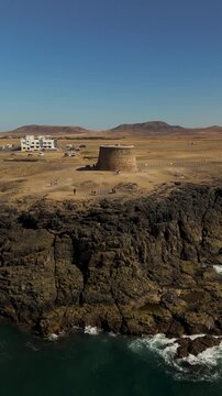 Dramatic vertical drone shot approaching the historic Torre del Tost&oacute;n fortress perched on the rugged volcanic cliffs of El Cotillo, Fuerteventura, Canary Islands.