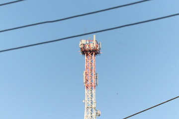 red and white telecommunications tower with antennas against blue sky seen through power lines