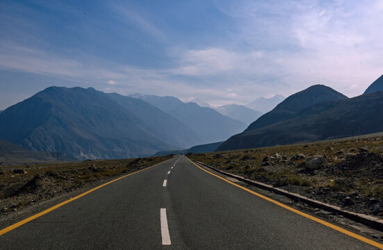 View of a long, straight asphalt road stretching into the distance towards hazy blue mountains under a bright sky, Jaglot, Gilgit Baltistan, Pakistan.