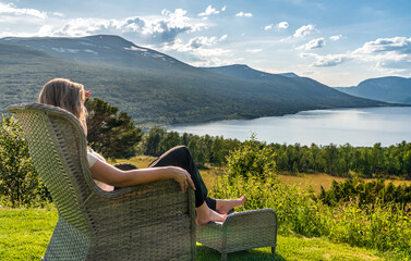 A woman sits in a garden chair with her feet up, looking at Gjevillvatnet lake and mountains in...