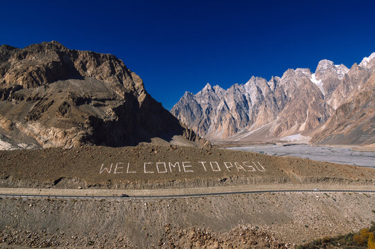 View of "WELCOME TO PASU" emblazoned on a hillside against the stark contrast of jagged, snow-capped peaks under a deep blue sky, Hunza Nagar, Gilgit Baltistan, Pakistan.