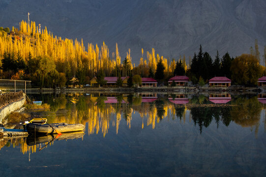 View of the golden trees reflecting in the calm water, creating a mirror image of the lakeside cabins and the distant, rugged mountains, Kachora, Gilgit Baltistan, Pakistan.