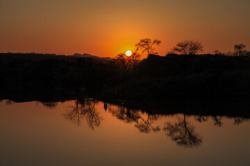 View of the sun's fiery orb descends beyond the horizon, casting a warm orange glow across still waters reflecting silhouetted trees, Chakwal, Punjab, Pakistan.