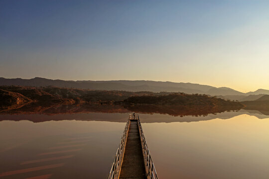 View of a solitary pier stretching into the still, reflective waters, mirroring the hazy hills under a gradient sky, bathed in the warm glow of dusk, Chakwal, Punjab, Pakistan.