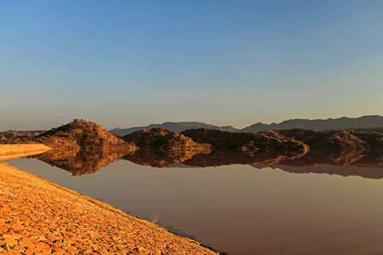 View of tranquil waters reflecting the rugged, golden hills under a serene blue sky, painting a picture of quiet beauty, Chakwal, Punjab, Pakistan.