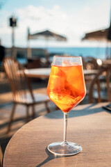 Aperol spritz cocktail with ice in a wine glass on a seaside cafe table. Refreshing summer aperitif with blurred beach background