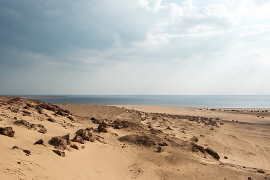 View of sun-baked dunes rolling towards the tranquil waters under a vast sky, a serene landscape painted in hues of gold and blue, Qaroun Protected Area, Faiyum Governorate, Egypt.