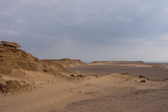 View of the arid golden sands and rocky plateaus meet the distant horizon under a cloudy sky, Qaroun Protected Area, Faiyum Governorate, Egypt.
