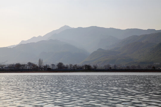 View of placid waters reflecting the soft light, meeting the distant shore where trees and structures lie beneath the hazy, layered mountains, Uchali Lake, Punjab, Pakistan.