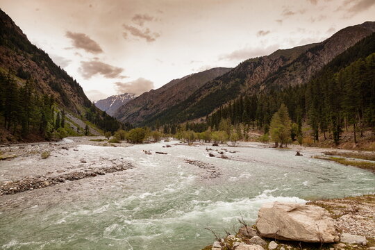 View of a vibrant river rushing through a valley, flanked by towering mountains and lush green trees under a cloudy sky, Kalam, Khyber Pakhtunkhwa, Pakistan.