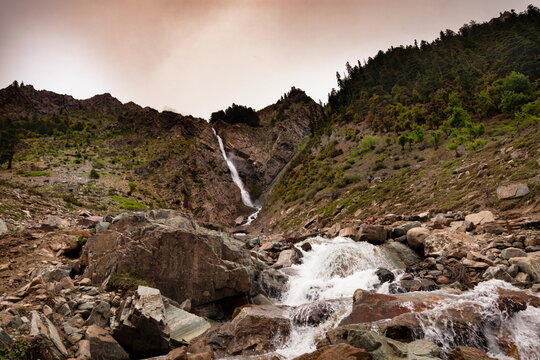View of rushing, foamy waters cascade over jagged rocks beneath the towering mountains and verdant slopes, creating a dynamic scene, Kalam, Khyber Pakhtunkhwa, Pakistan.