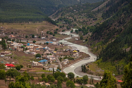 View of a meandering river carving through a valley dotted with small buildings and lush trees, framed by verdant mountains, Kalam, Khyber Pakhtunkhwa, Pakistan.