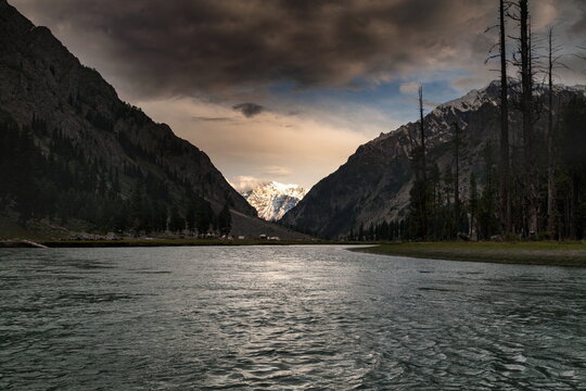 View of a serene river reflecting the ominous sky between towering mountains, one capped with snow, in Kalam, Khyber Pakhtunkhwa, Pakistan.