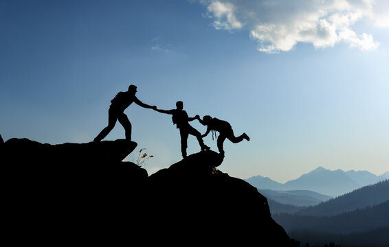 Silhouette of hikers helping each other climb a mountain peak at sunset, teamwork and collaboration concept.