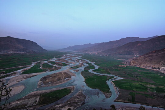 View of a braided river snaking its way through lush green fields, nestled between majestic mountains under a tranquil sky, Swat, Khyber Pakhtunkhwa, Pakistan.