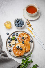 Pancakes with blueberry and banana on a plate on a light background with cup of tea